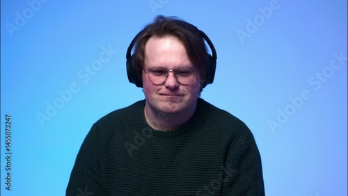 A portrait of a young light-brown-haired man with blue eyes, wearing glasses and headphones, listening to music and nodding his head to the rhythm.