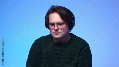 A portrait of a young light-brown-haired man with blue eyes, wearing glasses and headphones, listening to music and nodding his head to the rhythm.