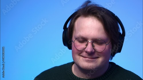 A portrait of a young light-brown-haired man with blue eyes, wearing glasses and headphones, listening to music and nodding his head to the rhythm.