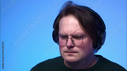 A portrait of a young light-brown-haired man with blue eyes, wearing glasses and headphones, listening to music and nodding his head to the rhythm.