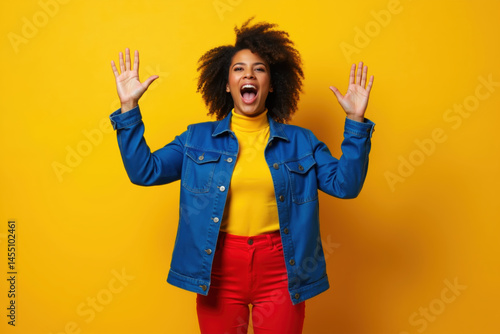 Obraz na płótnie A cheerful woman with curly hair, wearing a blue denim jacket, yellow turtleneck, and red pants, stands with arms raised against a vibrant yellow background. w salonie