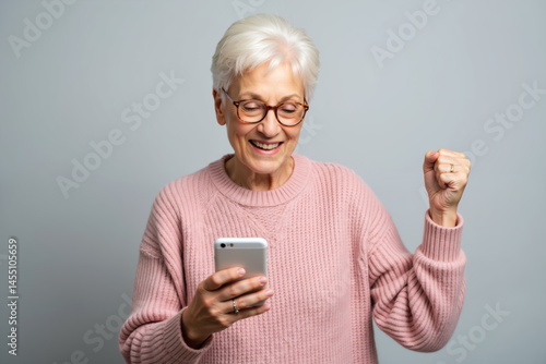 An elderly woman with white hair and glasses is happily looking at her smartphone while raising her fist in excitement, wearing a pink sweater.