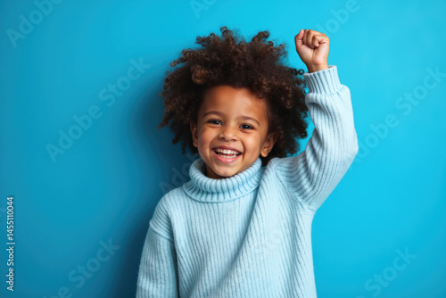 Obraz na płótnie A smiling child with curly hair, wearing a light blue sweater, stands against a blue background with a raised fist. w salonie