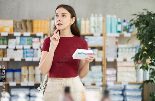 Woman choose buy paracetamol tablets box at pharmacy, compare with other good. Client check for absence of harmful substances, view product in details.