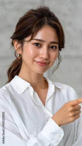 Attractive Asian woman points to the side with a friendly smile and wears a white collared shirt in a studio setting.