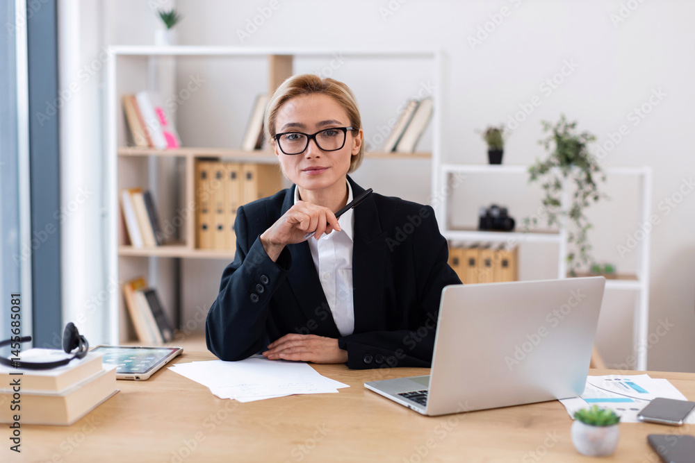 Fototapeta premium Confident adult woman in formal attire working in modern office environment. Professional focus with laptop, papers, books emphasizing business, professionalism, and productivity. Bright workspace.