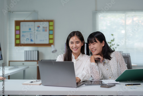 Two happy businesswomen are using a laptop and smiling while working together in a modern office, collaborating on a project and enjoying their teamwork