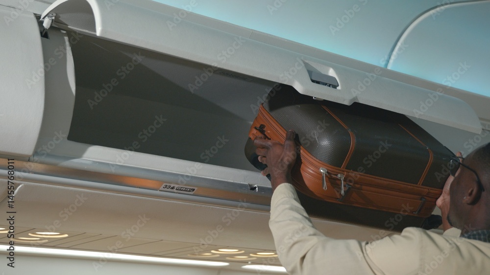 Naklejka premium Passenger black man putting luggage on top shelf or cabin compartment on airplane, business man lift suitcases putting overhead locker on airplane, Travel concept