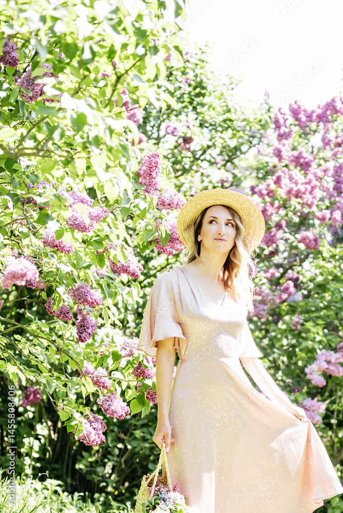 Fototapeta premium Young Woman In Summer Hat Enjoying The Fragrance Of Lilac Blooms On Sunny Day. lady in a straw hat and dress smelling lilac flowers in spring sunlit garden.