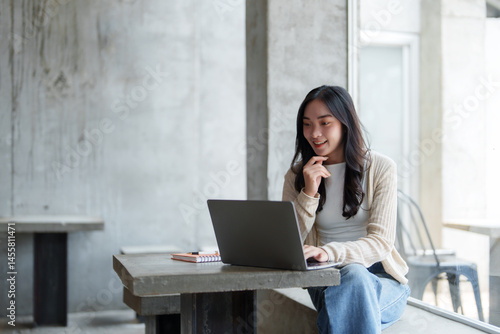Fotografi Smiling young asian woman sitting on a windowsill, working remotely on a laptop