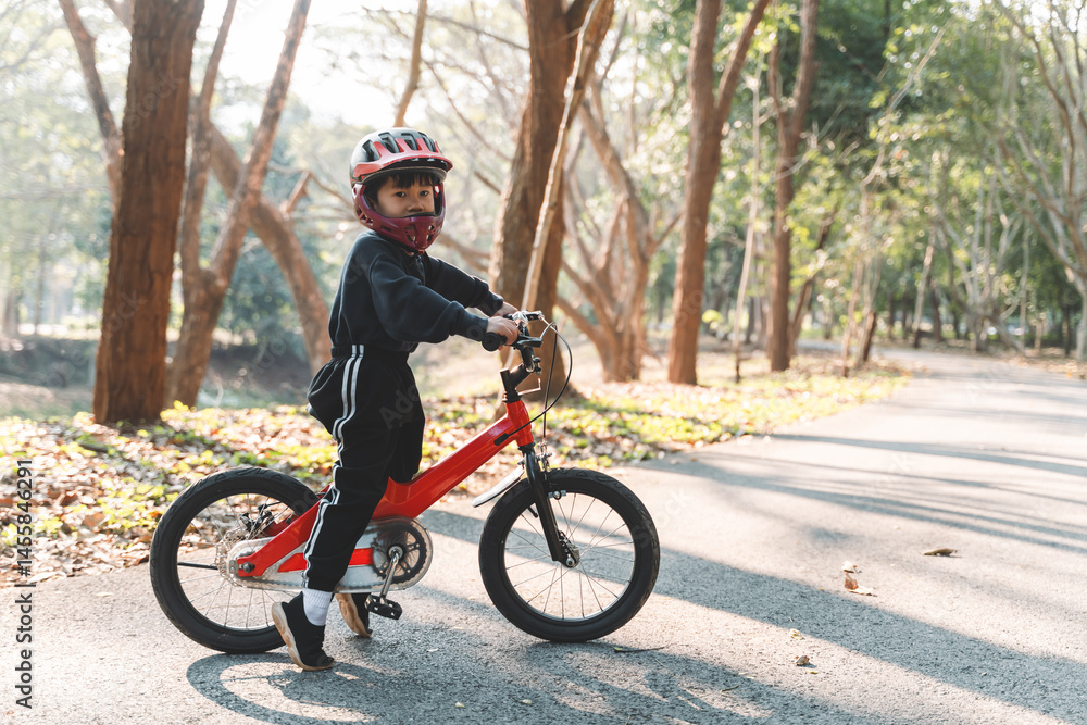 Fototapeta premium Young child with helmet learning to ride a red bicycle on a peaceful forest path in warm morning sunlight. Focused and determined expression.
