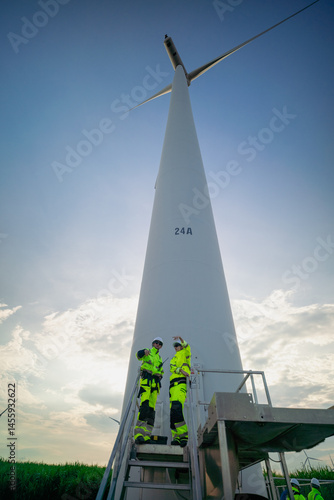 Maintenance engineer team standing at windmills at wind turbine farm. Group of people wear safety helmet and uniform working at alternative renewable energy wind station. Sustainable energy technology