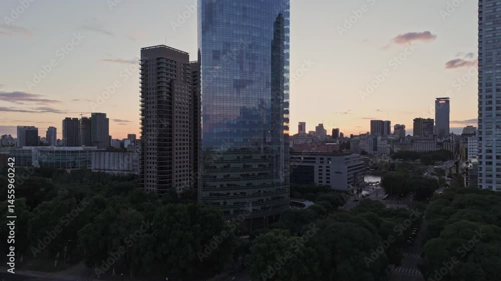 Smooth upward drone motion showcasing a tall reflective glass skyscraper surrounded by modern buildings in the Buenos Aires skyline. Captured during sunset with urban forest below. Smooth upward drone motion showcasing a tall reflective glass skyscraper surrounded by modern buildings in the Buenos Aires skyline. Captured during sunset with urban forest below.