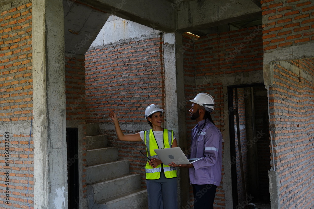 Black male engineer as foreman and Latin female construction inspector wearing construction helmet standing holding walkie talkie talking and consulting on construction of house project