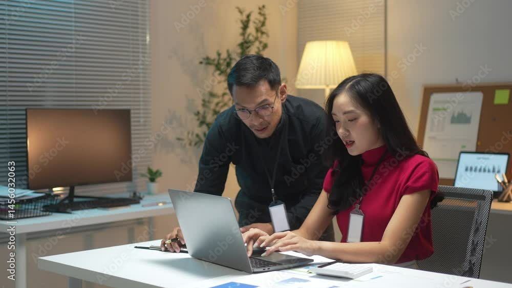 A man and a woman are sitting at a desk looking at a laptop. The man is smiling and the woman is smiling back. They seem to be working together on something