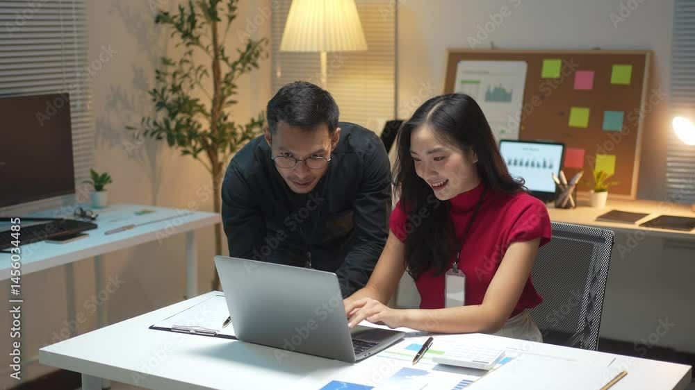 A man and a woman are sitting at a desk with a laptop open. The man is pointing at the screen while the woman looks on. Scene is one of collaboration and teamwork A man and a woman are sitting at a desk with a laptop open. The man is pointing at the screen while the woman looks on. Scene is one of collaboration and teamwork