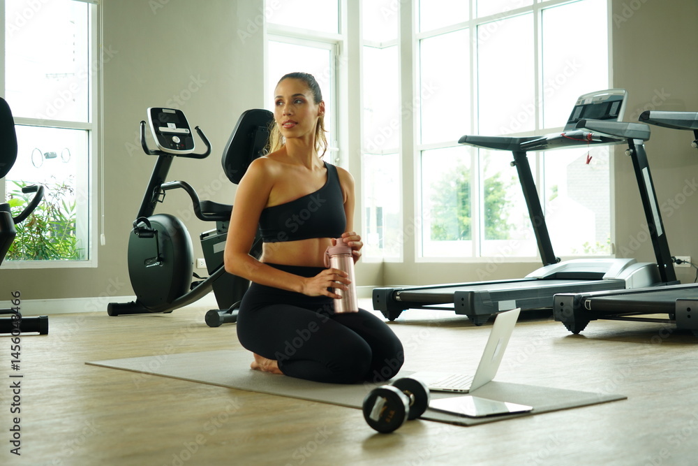 Beautiful Latin woman with a good figure sitting in a gym looking at a laptop to practice yoga exercise for health and weight loss in a fitness center. Health concept