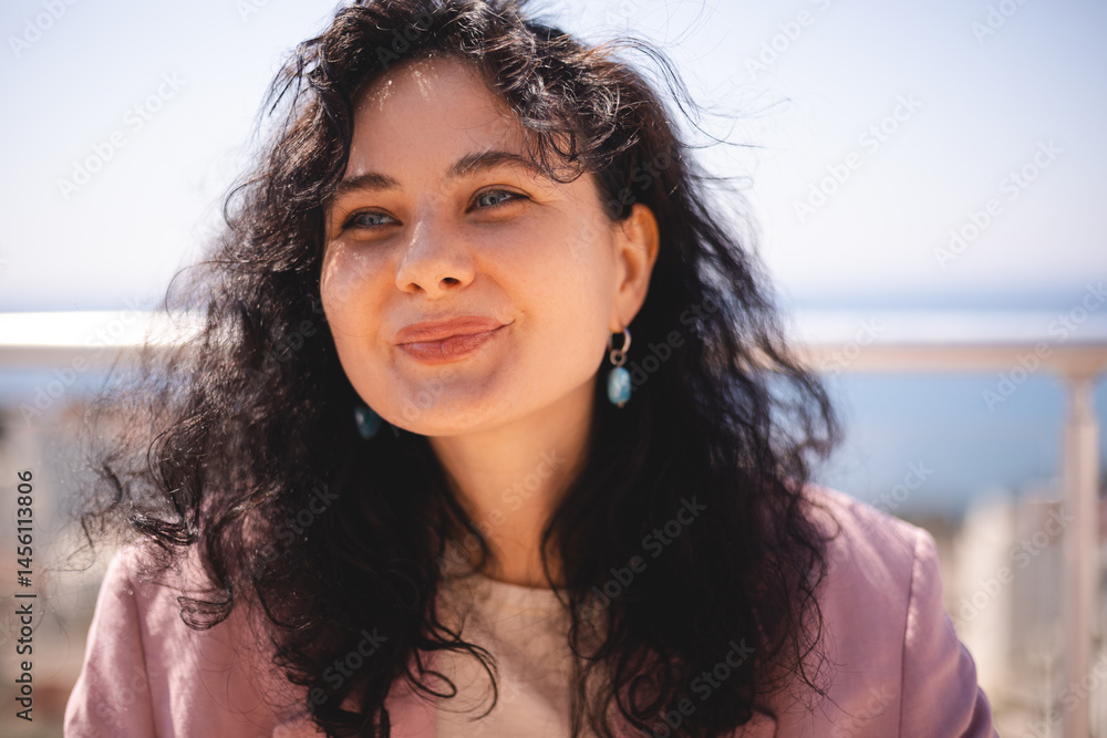 Fototapeta premium Charming brunette curly woman sitting on terrace near house, look at side, smiling, close up portrait. Girl wear pink jacket.