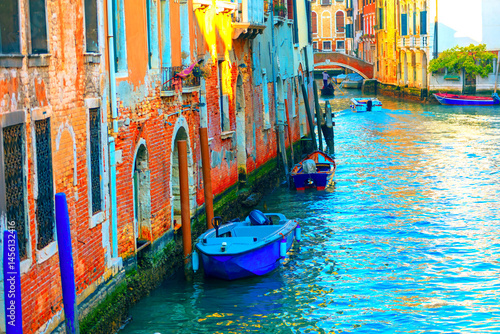 Narrow canal in Venice, flanked by weathered buildings with brick and plaster facades, illuminated by soft sunlight. Small boats tied to a pole, float on water, with stone bridge visible in background
