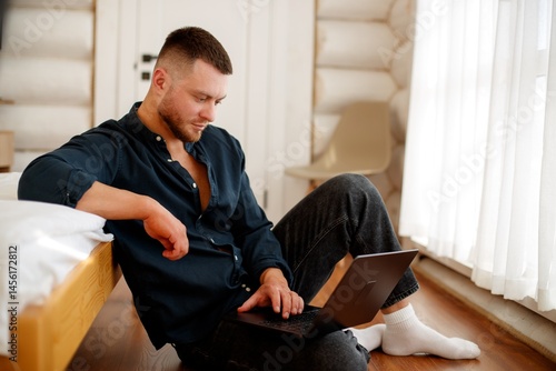 A man with a laptop in a cozy house. A man sits on the floor, focused on his laptop, surrounded by a cozy home environment with wooden walls.