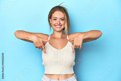 Blonde young woman in white top on blue background points down with fingers, positive feeling.