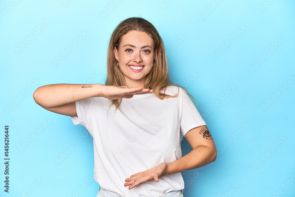 Fototapeta premium Blonde young woman in white t-shirt on blue background holding something with both hands, product presentation.