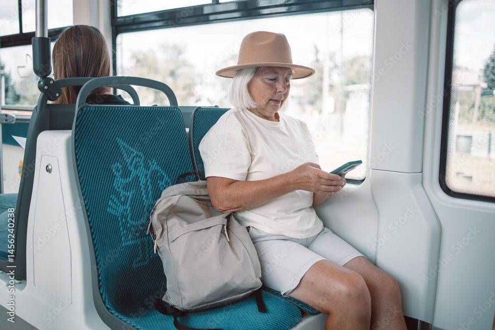 A senior woman wearing a pink hat is sitting on a bus, using her smartphone. She is traveling through the city, enjoying her journey in a relaxed manner.