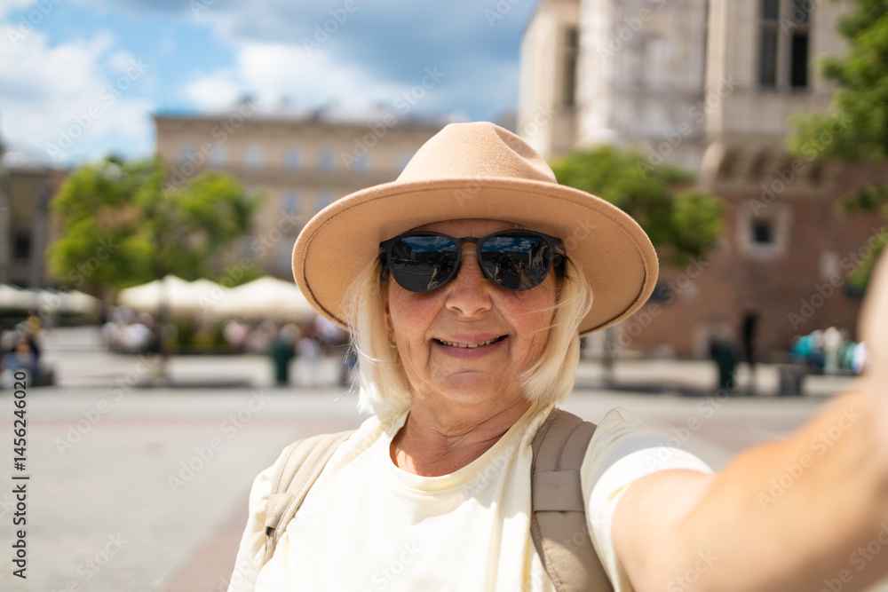 Senior woman smiles while taking a selfie in the city square, waving her hand. She is wearing a beige hat and sunglasses, enjoying a sunny day out while sightseeing.