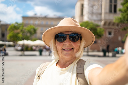 Obraz na płótnie Senior woman smiles while taking a selfie in the city square, waving her hand. She is wearing a beige hat and sunglasses, enjoying a sunny day out while sightseeing. w salonie