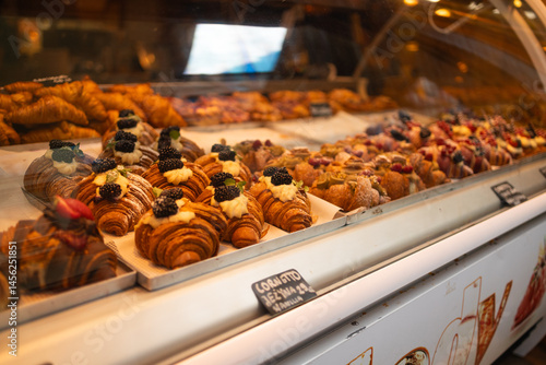 Obraz na płótnie A variety of fresh, golden croissants and pastries are displayed in a bakery showcase. The croissants are topped with colorful fruits and cream, inviting customers to indulge in these delicious treats w salonie