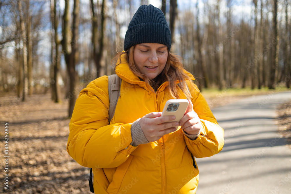 A happy woman in a yellow jacket and black beanie walking in the park, holding her phone. The bright natural setting, with trees and a peaceful path, enhances the cheerful mood of the moment.