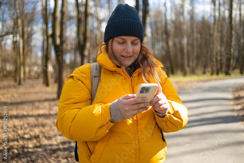 Obraz na płótnie A happy woman in a yellow jacket and black beanie walking in the park, holding her phone. The bright natural setting, with trees and a peaceful path, enhances the cheerful mood of the moment. w salonie