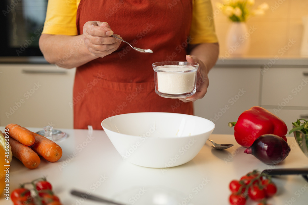 A senior woman in a yellow shirt and brown apron adds sugar to a bowl while preparing a meal in a well-organized kitchen. Fresh vegetables and kitchen tools are visible on the countertop