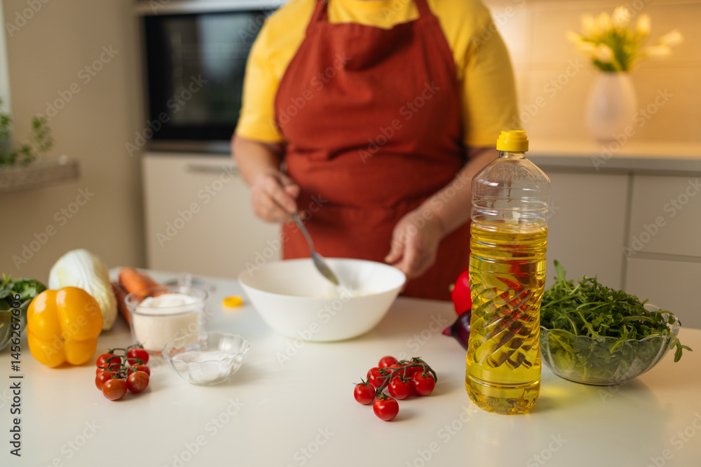A bottle of cooking oil stands on the kitchen counter, surrounded by fresh vegetables like tomatoes and greens, while a senior woman prepares a dish in the background