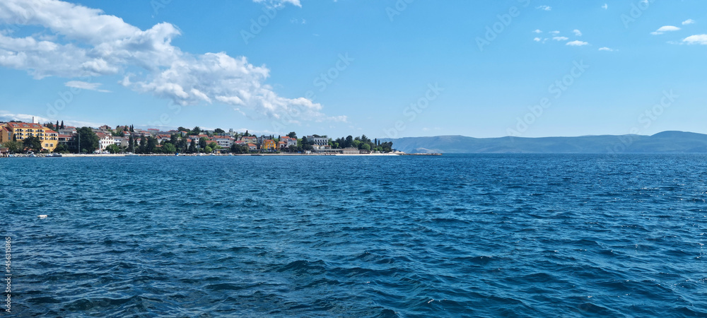 A picturesque coastal view of Selce, Croatia, shows colorful buildings lining the shore along the clear blue Adriatic Sea under a partly cloudy sky