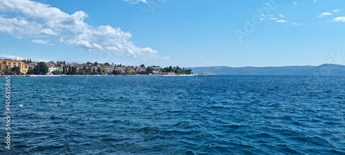 A picturesque coastal view of Selce, Croatia, shows colorful buildings lining the shore along the clear blue Adriatic Sea under a partly cloudy sky