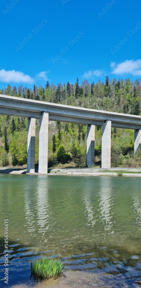 The Rijeka-Zagreb highway spans Lake Bajer on a concrete viaduct supported by tall pillars, with forested hills in the background under a blue sky with some clouds