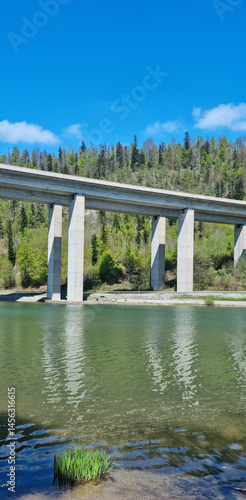 The Rijeka-Zagreb highway spans Lake Bajer on a concrete viaduct supported by tall pillars, with forested hills in the background under a blue sky with some clouds