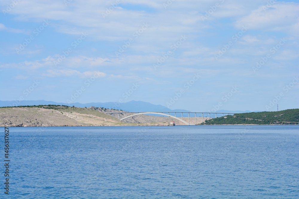 A scenic view from Jadranovo captures the Krk Bridge spanning the blue sea, connecting the mainland to Krk Island under a partly cloudy sky
