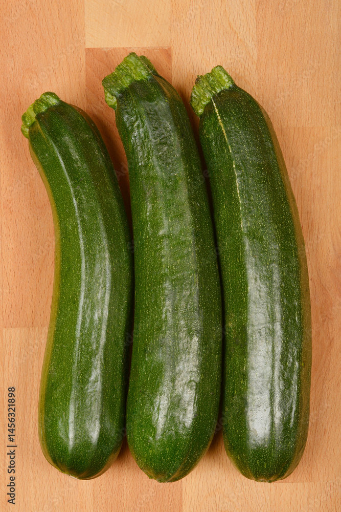 Three green zucchinis are arranged vertically on a light brown wooden surface