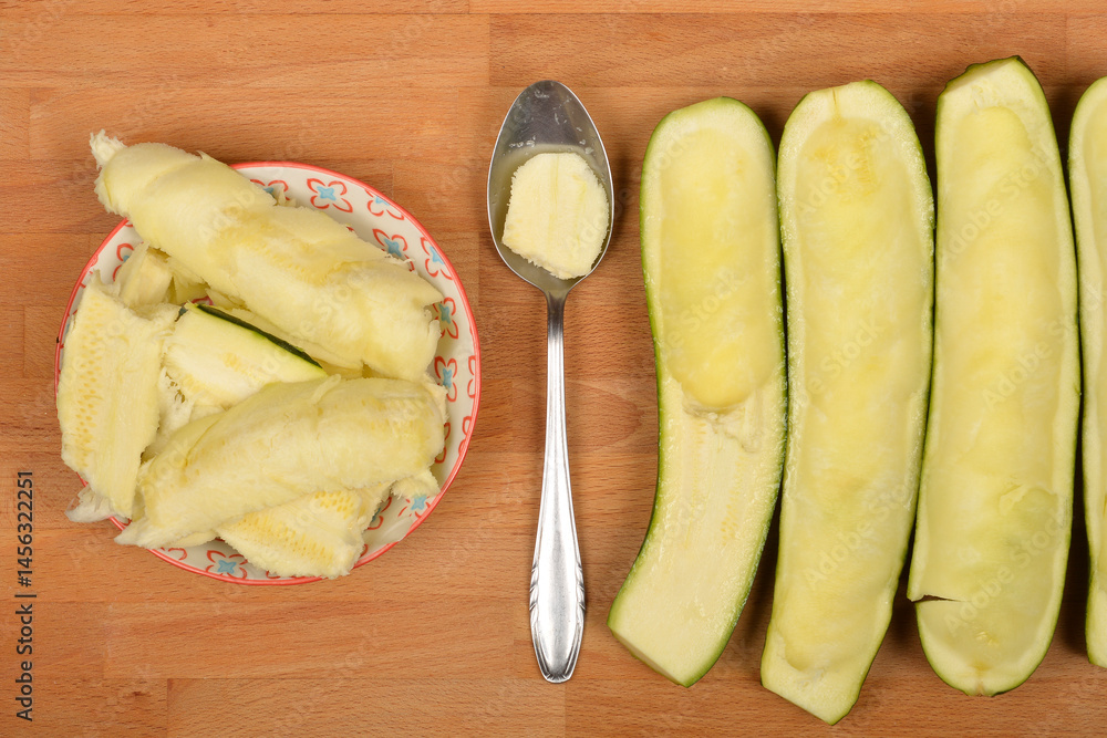 Halved and scooped-out zucchinis are arranged with removed pulp in a bowl and on a spoon on a wooden board