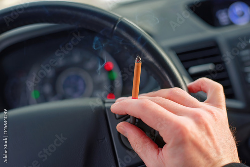 Hand holding burning cigarette while gripping steering wheel, with smoke filling car, distracted driving and vehicle malfunction. Unhealthy habits, and emergency roadside situations. Selective focus