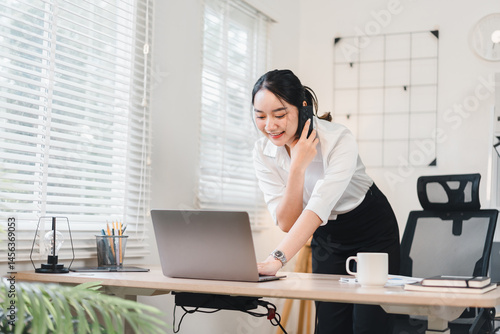 Young Asian businesswoman standing at modern office desk, smiling while talking on phone and working on laptop, bright workspace with natural light, professional and productive atmosphere