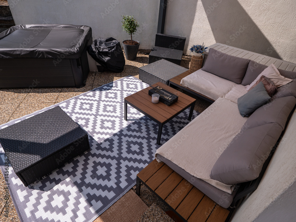 Fototapeta premium Modern aluminum patio with outdoor sofa, hot tub, coffee tables, and decorative rug on stone tiles, partially shaded, with potted plant and wall in the background.