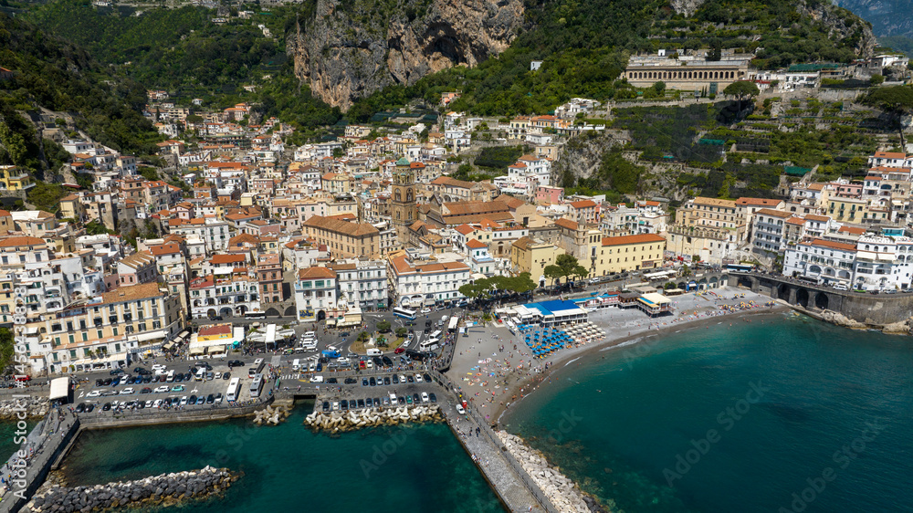 Obraz premium Aerial view of the historic center of Amalfi, in the province of Salerno, in Campania, Italy. Among the alleys there's the Cathedral of the town. In foreground is the beach with umbrellas and tourists