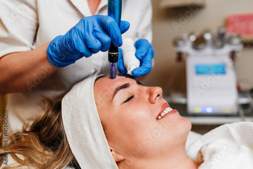 Beautician wearing blue gloves uses a microneedling pen to apply serum to forehead of client with closed eyes lying on a treatment table during a beauty spa procedure. Modern anti-age procedure.