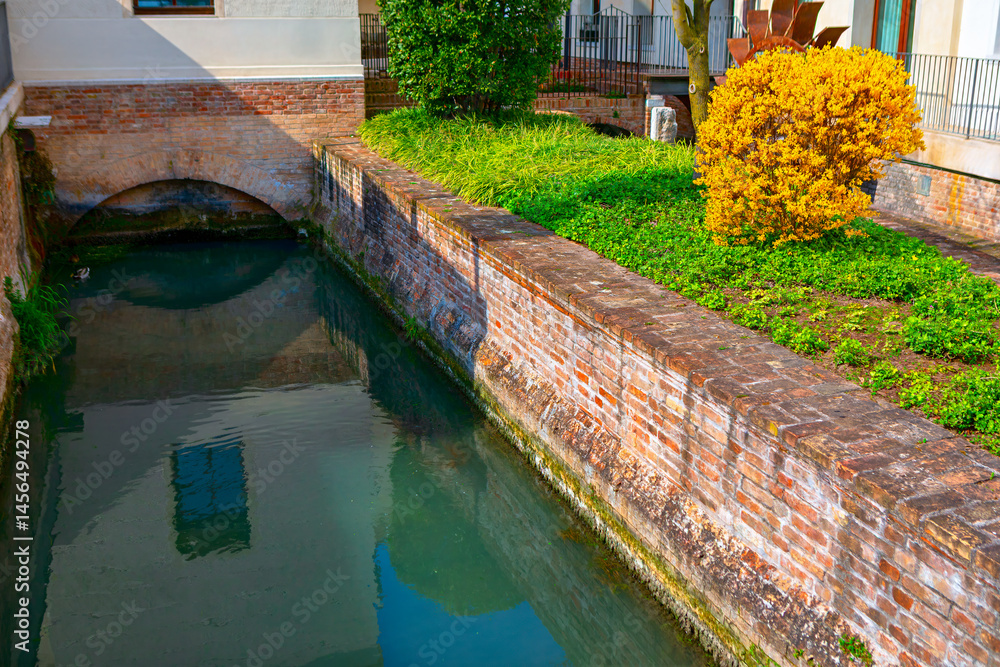 Narrow canal bordered by tall brick walls, with calm water reflecting surroundings. Lush green grass and a vibrant yellow shrub add color to scene, with a small arched bridge visible in background