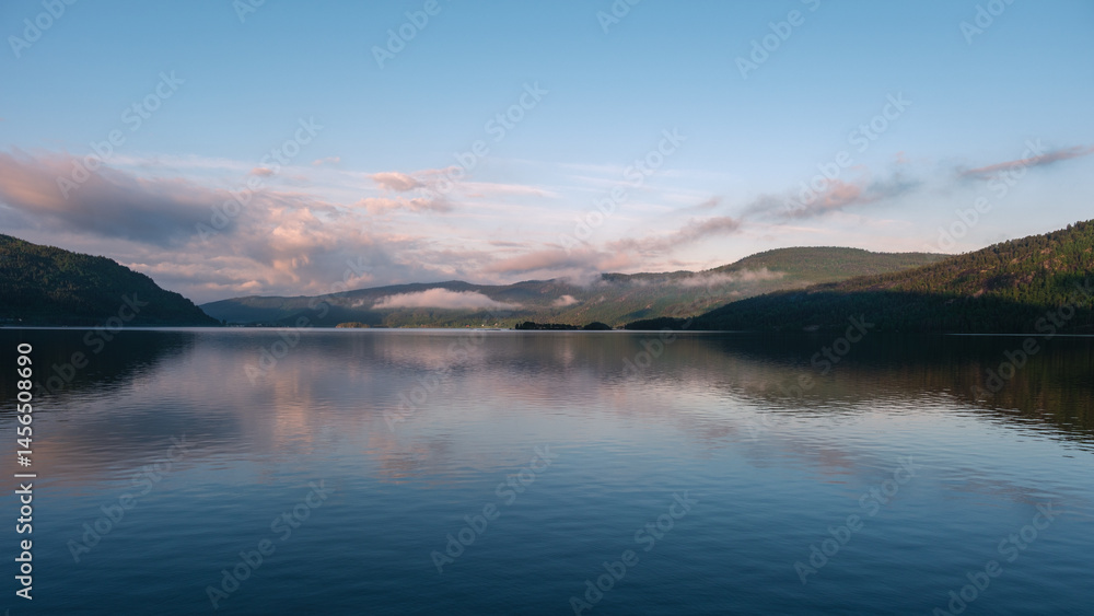 Fototapeta premium Morning light casts a peaceful glow over the tranquil waters of a lake in Norway. Mist gently rises from the surrounding hills, creating a serene atmosphere perfect for relaxation and inspiration.