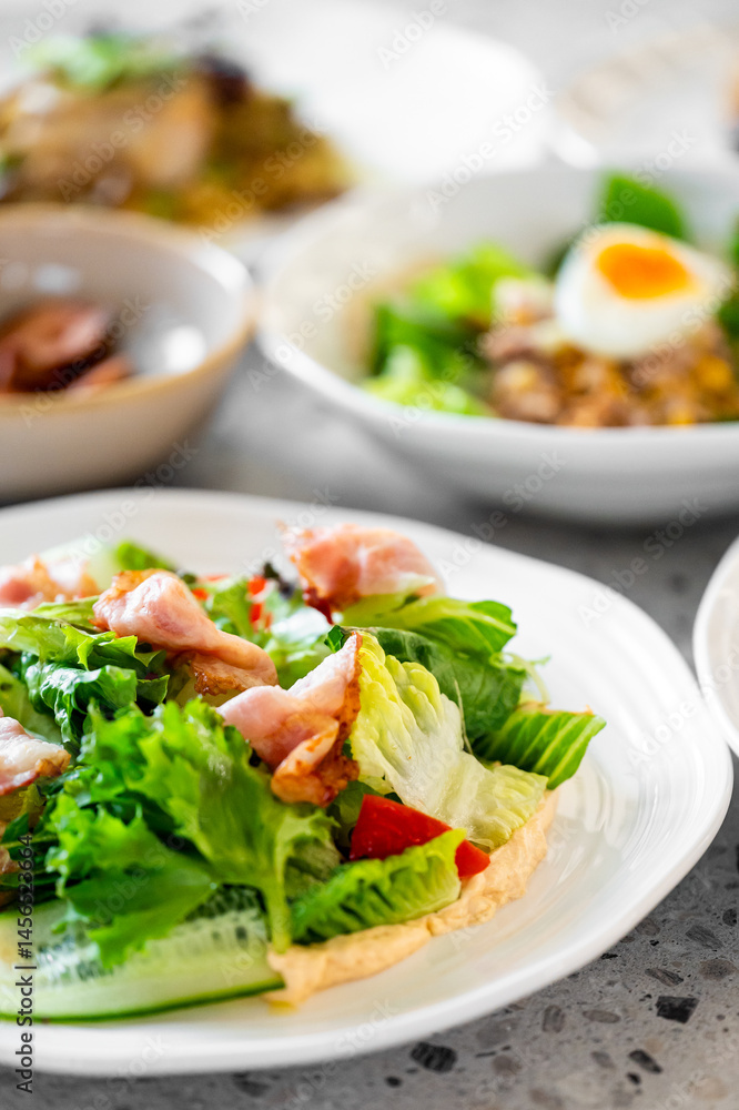Fresh salad with crispy bacon, lettuce, cucumber, and red bell pepper on a white plate. A bowl with a boiled egg and greens is visible in the background. Vibrant colors and healthy ingredients. Fresh salad with crispy bacon, lettuce, cucumber, and red bell pepper on a white plate. A bowl with a boiled egg and greens is visible in the background. Vibrant colors and healthy ingredients.