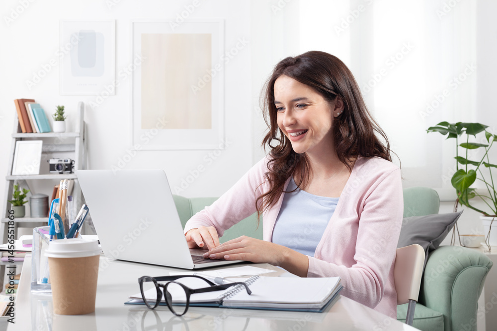Happy young woman at home, sitting at a cozy desk and smiling at her laptop. Representing a positive remote work or online learning, video call, or digital collaboration in comfortable home setting Happy young woman at home, sitting at a cozy desk and smiling at her laptop. Representing a positive remote work or online learning, video call, or digital collaboration in comfortable home setting